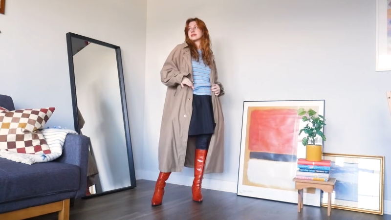 Devojka u mini suknji, dugom bež kaputu i crvenim čizmama pozira u dnevnoj sobi