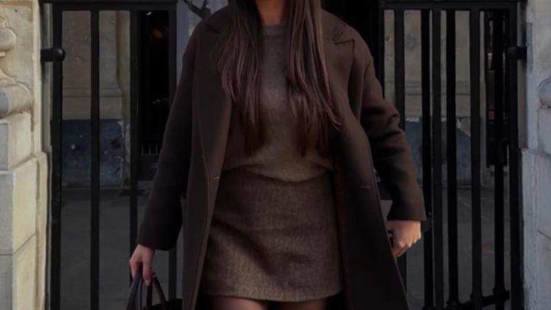 Žena u braon mini suknji i dugom kaputu stoji ispred crne kapije i drži torbu u ruci