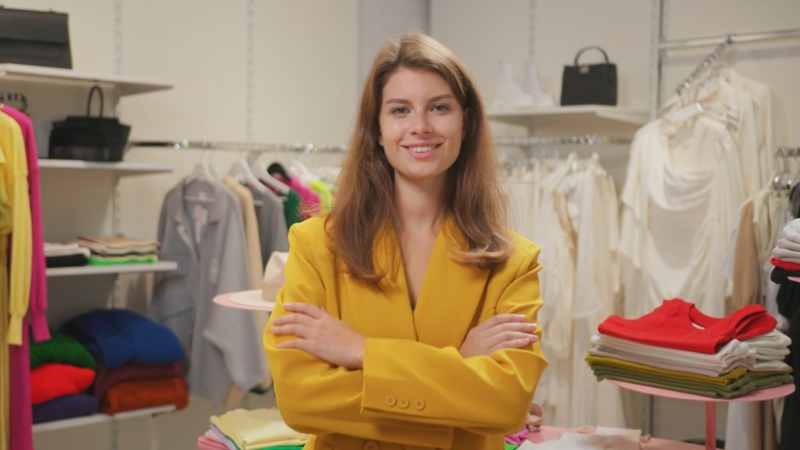 Žena u žutom blejzeru stoji u butiku sa prekrštenim rukama